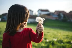 Copil ținând o păpădie în mână, imagine simbolică pentru diferențele dintre copii și unicitatea fiecărui mod de dezvoltare.